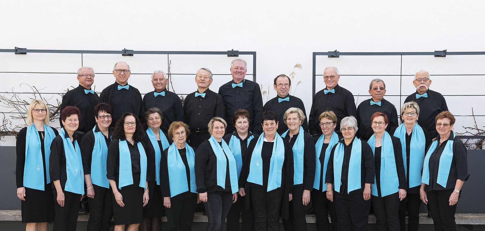 Gruppe von 21 Personen, dem Liederkranz Monheim, in schwarzer Kleidung mit türkisfarbenen Schals und Fliegen, posiert vor einer weißen Wand auf einem gepflastertem Platz.