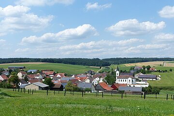 Aufnahme des kleines Dorfes Wittesheim mit roten Dächern und einer Kirche, umgeben von grünen Feldern und Wäldern unter blauem Himmel mit Wolken. Vorderhalb ein eingezäunter Bereich.