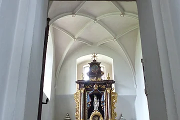 Ein Blick durch eine Tür zu einem Kirchenaltar in der Walburgakapelle in Monheim, mit goldenen Verzierungen und einer Statue, der heiligen Walburga. Der Kirchenaltar ist umgeben von Kerzen und Blumen, in einem gewölbten Raum mit einer verzierten Decke.