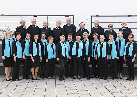 Gruppe von 21 Personen, dem Liederkranz Monheim, in schwarzer Kleidung mit türkisfarbenen Schals und Fliegen, posiert vor einer weißen Wand auf einem gepflastertem Platz.