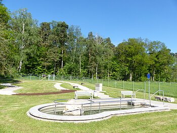 Rundes Kneippbecken in Monheim mit einem Geländer und einem Barfußpfad auf einer Wiese, umgeben von einer großen Grünfläche und Bäumen und Sitzmöglichkeiten. Ein Zaun im Hintergrund.