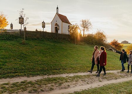 Vier Personen spazieren auf einem Weg an einer kleinen Kapelle vorbei, der Kalvarienbergkapelle in Wittesheim, umgeben von herbstlicher Landschaft. Auf dem Weg vor der Kapelle stehen Kreuzwegstationen.
