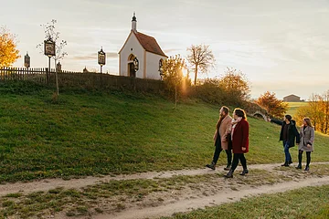 Vier Personen spazieren auf einem Weg an einer kleinen Kapelle vorbei, der Kalvarienbergkapelle in Wittesheim, umgeben von herbstlicher Landschaft. Auf dem Weg vor der Kapelle stehen Kreuzwegstationen.