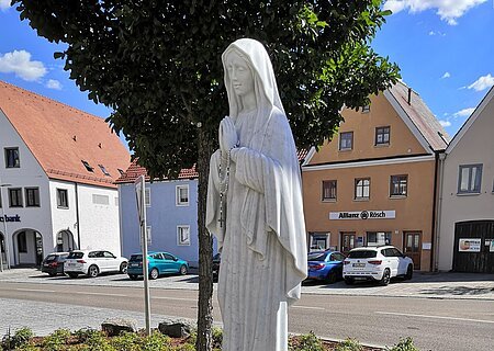 Statue einer betenden Frau mit Rosenkranz, die Marienstatue auf einem kleinen Podest, umgeben von Blumen, im Hintergrund eine Straße und ein Parkplatz mit Gebäuden.