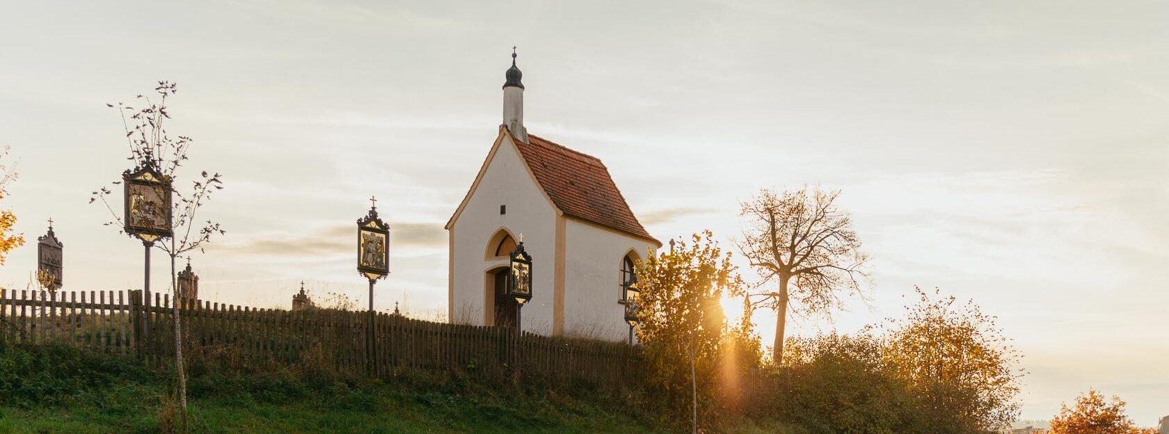 Vier Personen spazieren auf einem Weg an einer kleinen Kapelle vorbei, der Kalvarienbergkapelle in Wittesheim, umgeben von herbstlicher Landschaft. Auf dem Weg vor der Kapelle stehen Kreuzwegstationen.