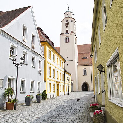 Gepflasterte Gasse mit bunten Häusern und einem Kirchturm im Hintergrund bei Tageslicht.