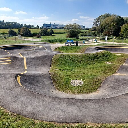 Asphaltierter Pumptrack mit gelben Markierungen in der Mitte, umgeben von grüner Landschaft und Bäumen im Hintergrund.