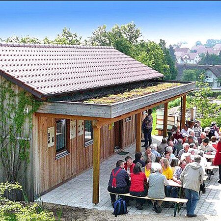 Holzgebäude mit einer Terrasse, davor sitzen und stehen Menschen in Gruppen, im Hintergrund eine grüne Landschaft mit Häusern.