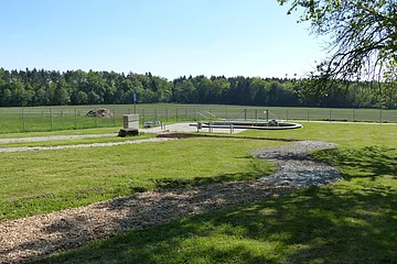 Grüne Wiese mit einem geschwungenem Barfußpfad mit Holzschnitzeln und Steinen und umzäuntem Bereich im Hintergrund, umgeben von Bäumen.