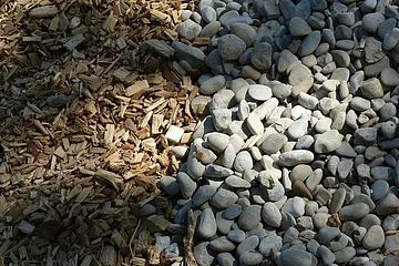 Holzhackschnitzel links neben einer Fläche mit kleinen, grauen Steinen von der Sonne bestrahlt.