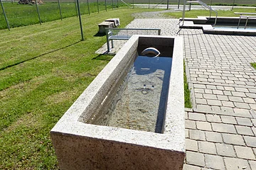 Steinbecken der Kneippanlage in Monheim mit klarem Wasser auf gepflastertem Platz, im Hintergrund Wiese und Zaun.