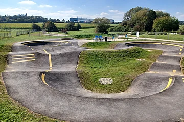 Asphaltierter Pumptrack mit gelben Markierungen in der Mitte, umgeben von grüner Landschaft und Bäumen im Hintergrund.