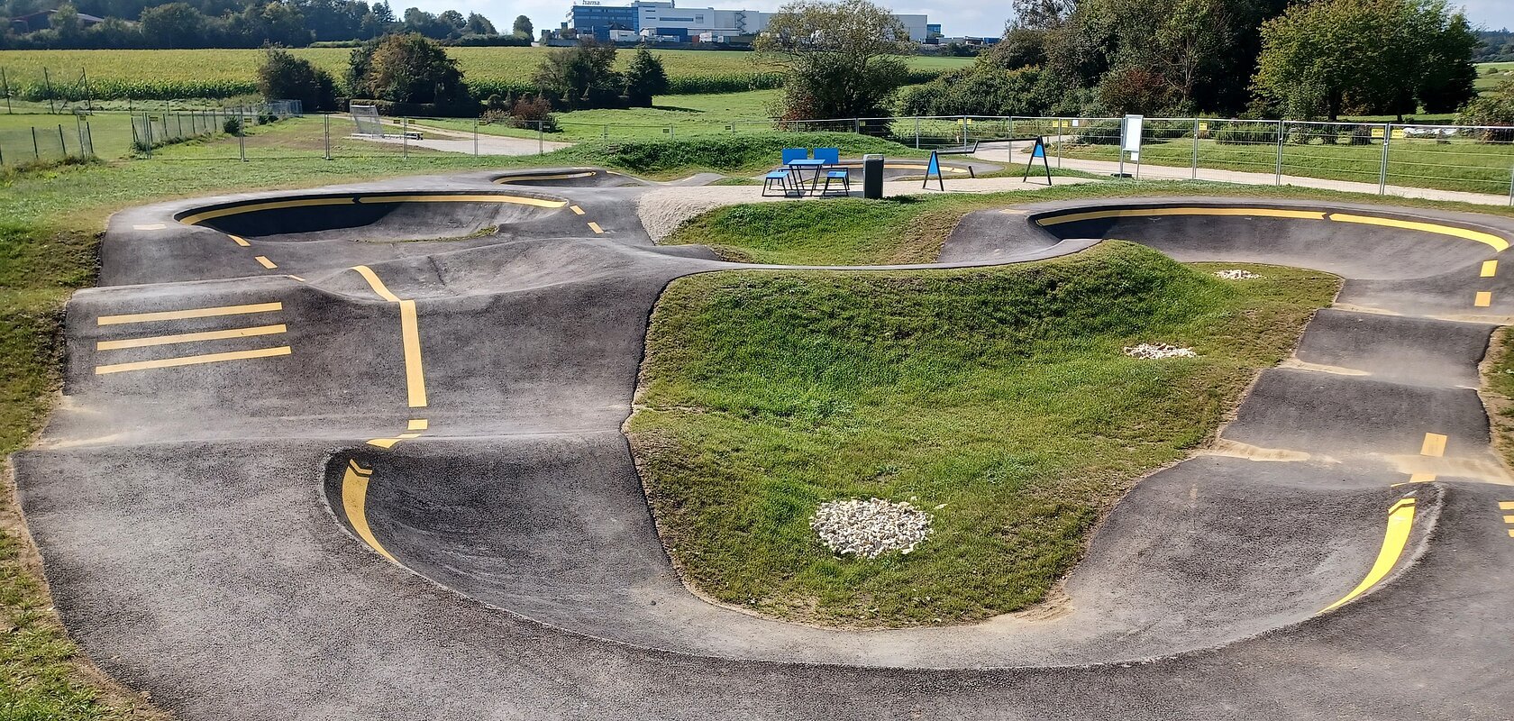 Asphaltierter Pumptrack mit gelben Markierungen in der Mitte, umgeben von grüner Landschaft und Bäumen im Hintergrund.