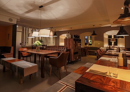 Ein Restaurant mit Holztischen, Stühlen und gedämpfter Beleuchtung. Auf den Tischen Besteck und Gewürzbehälter und auf einem Tisch stehen Blumen in der Vase.