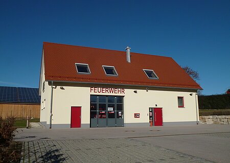 Gelbes Feuerwehrgebäude mit rotem Dach, Aufschrift "Feuerwehr" an der Fassade, zwei rote Türen, ein großes Tor und Fenster auf der Vorderseite. Vorderhalb gepflasterter Platz und blauer Himmel im Hintergrund.