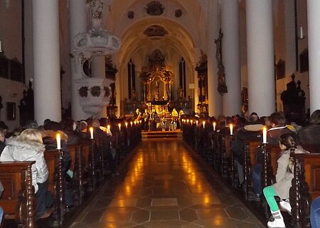 Innenraum einer Kirche mit Kerzen beleuchtet, Menschen sitzen auf Holzbänken, Blick auf den Altar im Hintergrund.
