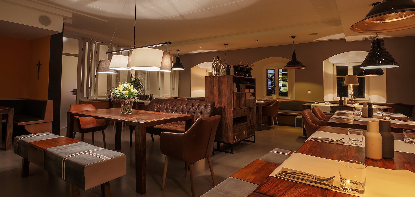 Ein Restaurant mit Holztischen, Stühlen und gedämpfter Beleuchtung. Auf den Tischen Besteck und Gewürzbehälter und auf einem Tisch stehen Blumen in der Vase.