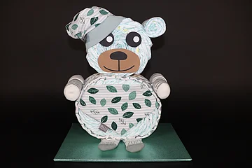 Windeltorte in Form eines Bären mit Blättermuster, trägt Mütze und Socken, steht auf grünem Untergrund vor schwarzem Hintergrund.