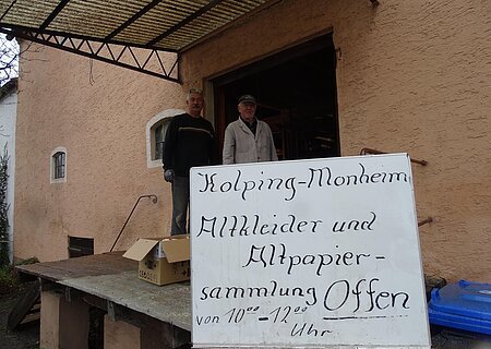 Zwei Personen stehen vor einem orangegestrichenen Gebäude neben einem Schild mit der Aufschrift "Kolping-Monheim Altkleider und Altpapier-Sammlung  Offen".