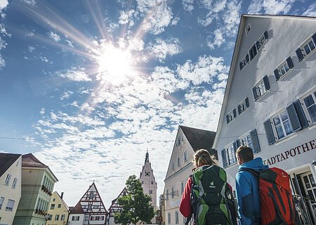 Zwei Personen mit Rucksäcken gehen durch eine Stadt mit Fachwerkhäusern unter einem sonnigen Himmel. Auf dem Haus im Vordergrund steht die Aufschrift "Stadtapotheke".