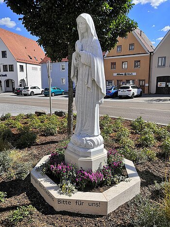Marienstatue "Maria, Königin des Friedens" Statue einer betenden Frau mit Rosenkranz, die Marienstatue auf einem kleinen Podest, umgeben von Blumen, im Hintergrund eine Straße und ein Parkplatz mit Gebäuden.