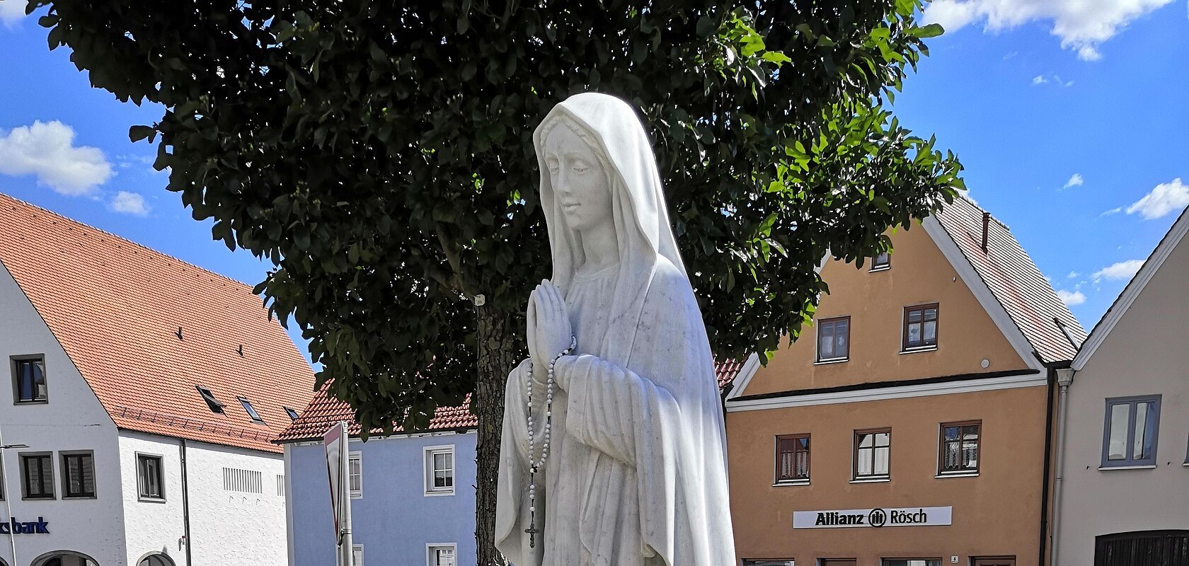 Statue einer betenden Frau mit Rosenkranz, die Marienstatue auf einem kleinen Podest, umgeben von Blumen, im Hintergrund eine Straße und ein Parkplatz mit Gebäuden.
