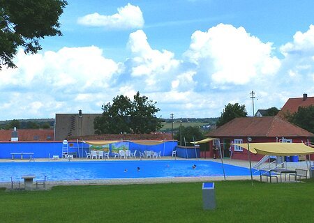 Freibad mit blauem Wasser, umgeben von Rasen, Bäumen und Gebäuden unter einem bewölkten Himmel. Vorderhalb vom Becken sind Sitzmöglichkeiten.