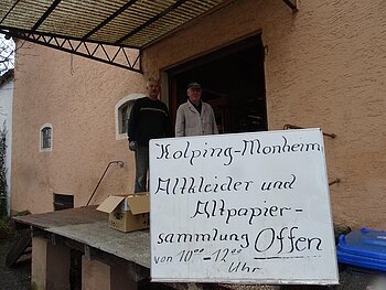 Zwei Personen stehen vor einem orangegestrichenen Gebäude neben einem Schild mit der Aufschrift "Kolping-Monheim Altkleider und Altpapier-Sammlung  Offen".
