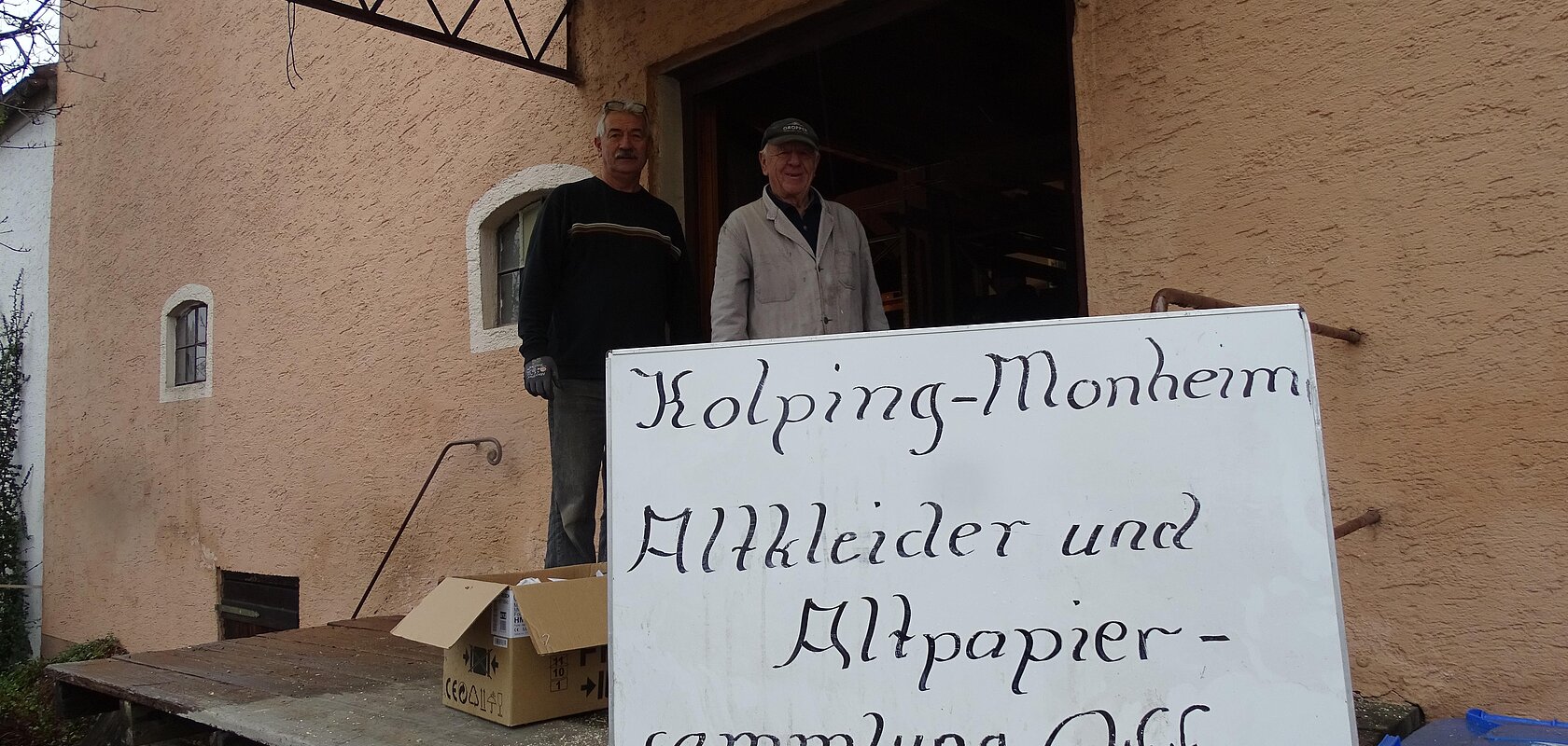 Zwei Personen stehen vor einem orangegestrichenen Gebäude neben einem Schild mit der Aufschrift "Kolping-Monheim Altkleider und Altpapier-Sammlung  Offen".