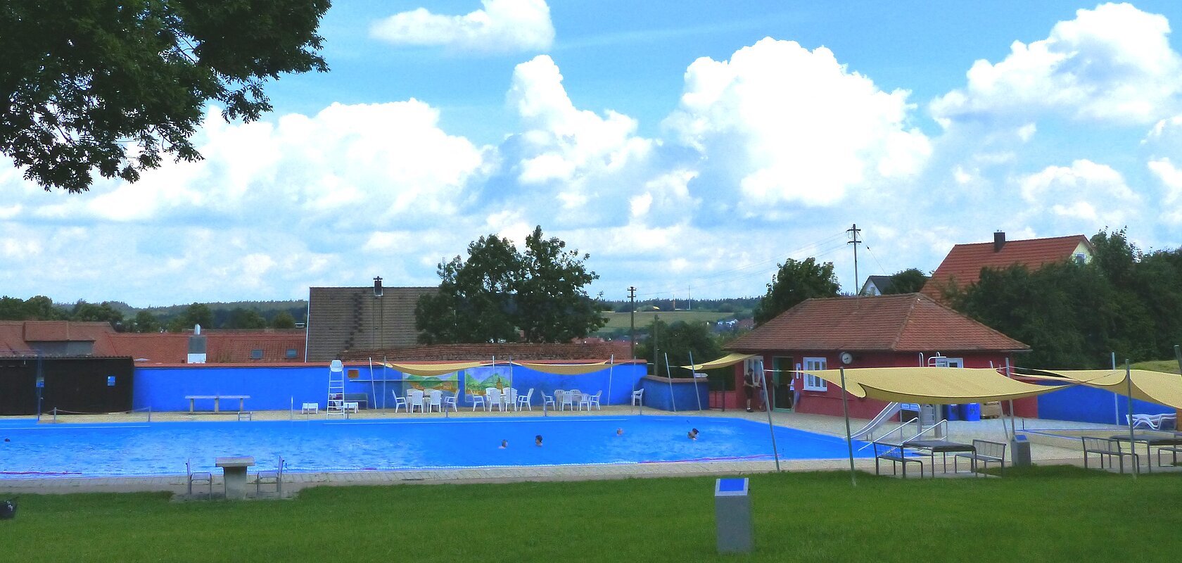 Freibad mit blauem Wasser, umgeben von Rasen, Bäumen und Gebäuden unter einem bewölkten Himmel. Vorderhalb vom Becken sind Sitzmöglichkeiten.