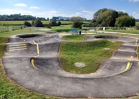 Asphaltierter Pumptrack mit gelben Markierungen in der Mitte, umgeben von grüner Landschaft und Bäumen im Hintergrund.