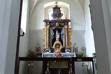 Ein Blick durch eine Tür zu einem Kirchenaltar in der Walburgakapelle in Monheim, mit goldenen Verzierungen und einer Statue, der heiligen Walburga. Der Kirchenaltar ist umgeben von Kerzen und Blumen, in einem gewölbten Raum mit einer verzierten Decke.