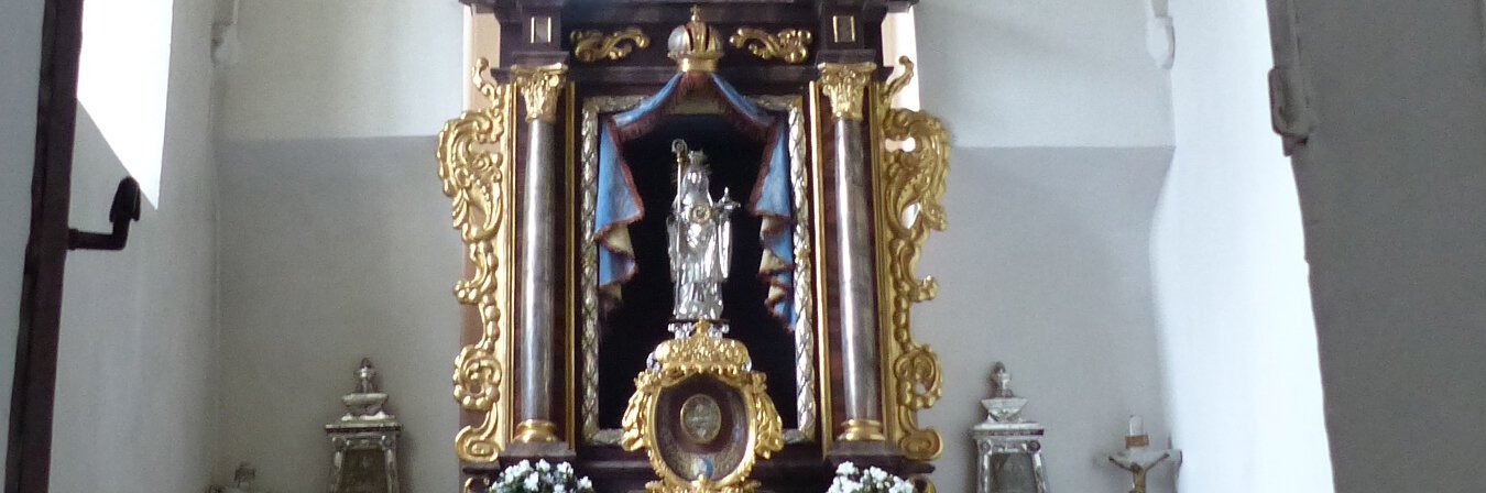 Ein Blick durch eine Tür zu einem Kirchenaltar in der Walburgakapelle in Monheim, mit goldenen Verzierungen und einer Statue, der heiligen Walburga. Der Kirchenaltar ist umgeben von Kerzen und Blumen, in einem gewölbten Raum mit einer verzierten Decke.