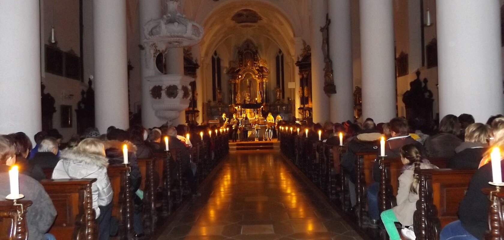 Innenraum einer Kirche mit Kerzen beleuchtet, Menschen sitzen auf Holzbänken, Blick auf den Altar im Hintergrund.