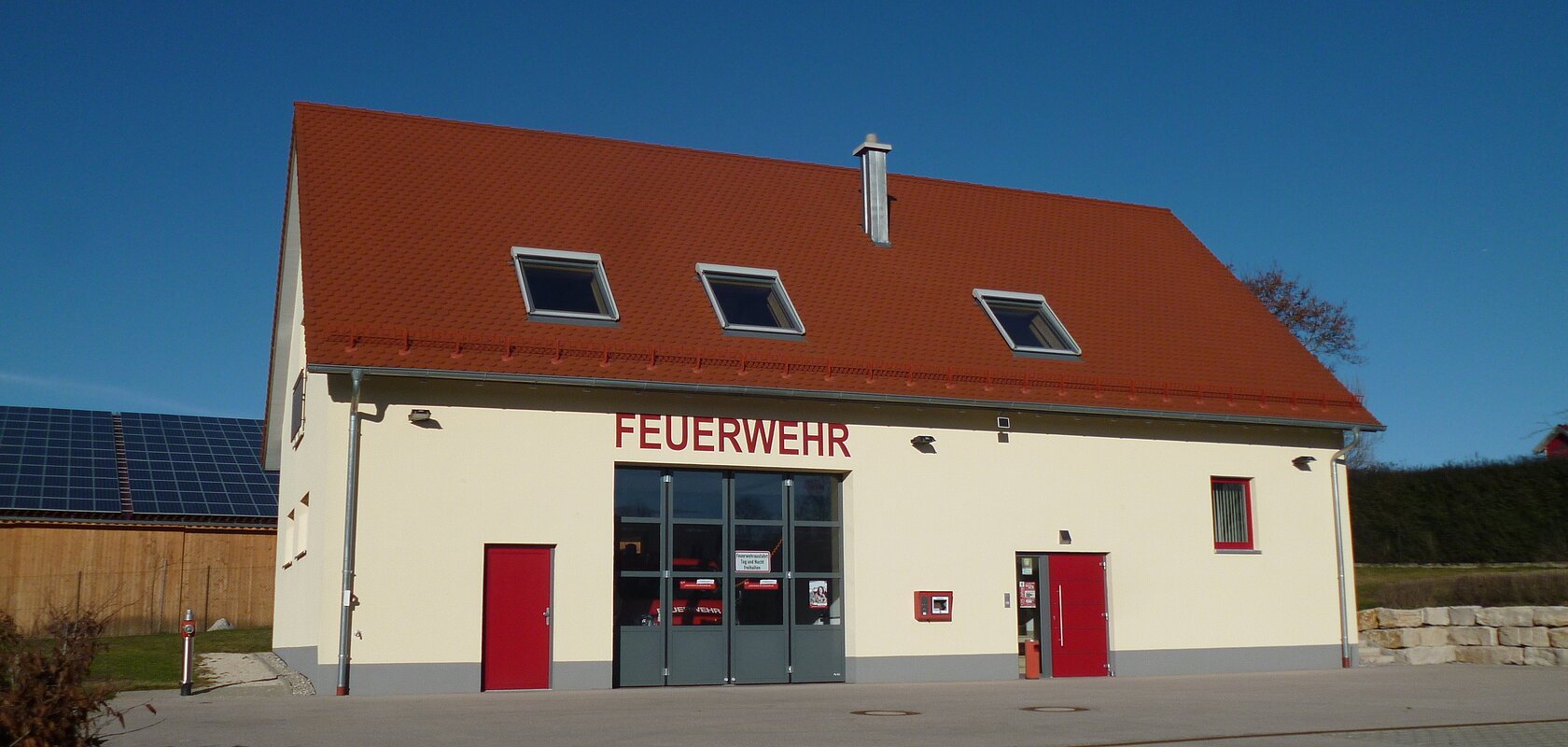 Gelbes Feuerwehrgebäude mit rotem Dach, Aufschrift "Feuerwehr" an der Fassade, zwei rote Türen, ein großes Tor und Fenster auf der Vorderseite. Vorderhalb gepflasterter Platz und blauer Himmel im Hintergrund.