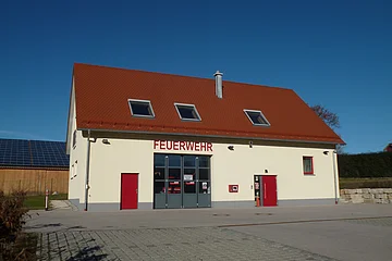 Gelbes Feuerwehrgebäude mit rotem Dach, Aufschrift "Feuerwehr" an der Fassade, zwei rote Türen, ein großes Tor und Fenster auf der Vorderseite. Vorderhalb gepflasterter Platz und blauer Himmel im Hintergrund.