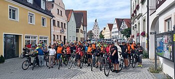 Gruppe von Radfahrern mit Helmen und orangefarbenen Trikots auf einer gepflasterten Straße in einer Altstadt mit Fachwerkhäusern bei blauem Himmel. Vorne rechts im Bild ein Schaukasten mit Aushängen.