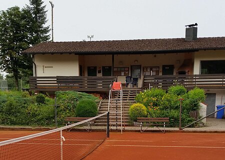 Tennisplatz mit rotem Sandbelag, einem Netz in der Mitte, dahinter ein Gebäude mit braunem Dach, einer Terrasse und Treppe, umgeben von Büschen und Bäumen.