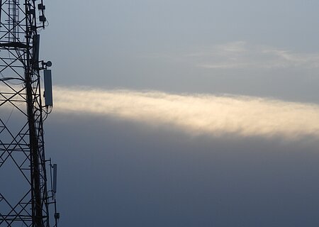 Mobilfunk-Sendemast vor bewölktem Himmel mit einer schmalen, hellen Wolke im Hintergrund.