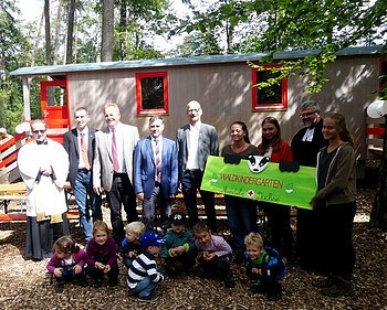Waldkindergarten Mandele Dachse in Monheim eröffnet Eine Gruppe von Erwachsenen und Kindern steht bzw. kniet vor einem Bauwagen im Wald, drei Personen halten ein gestaltetes Schild mit der Aufschrift "Waldkindergarten Mandele Dachse" in den Händen.