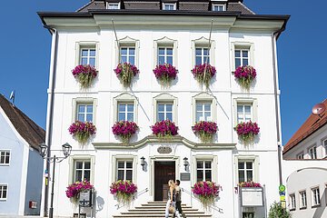 Das Rathaus in Monheim: Ein weißes dreistöckiges Gebäude mit mehreren Fenstern, an denen Blumenkästen mit pinken Blumen hängen. Zwei Personen gehen davor vorbei. Links und rechts davon jeweils ein weiteres Haus.