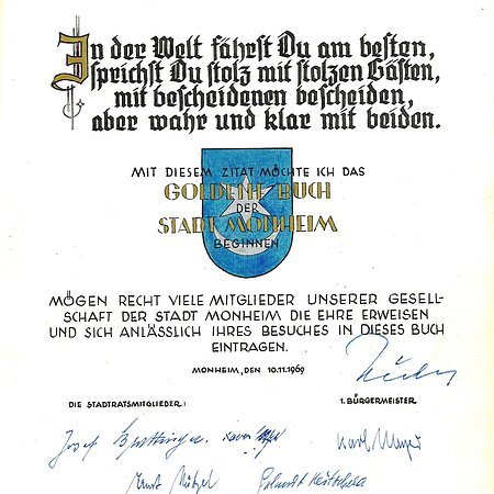 Erster Eintrag im Goldenen Buch der Stadt Monheim: Oben ein Zitat, darunter das Wappen der Stadt Monheim in blau. Darunter ein weiterer Text, datiert mit 10.11.1969, mit etlichen Unterschriften darunter.