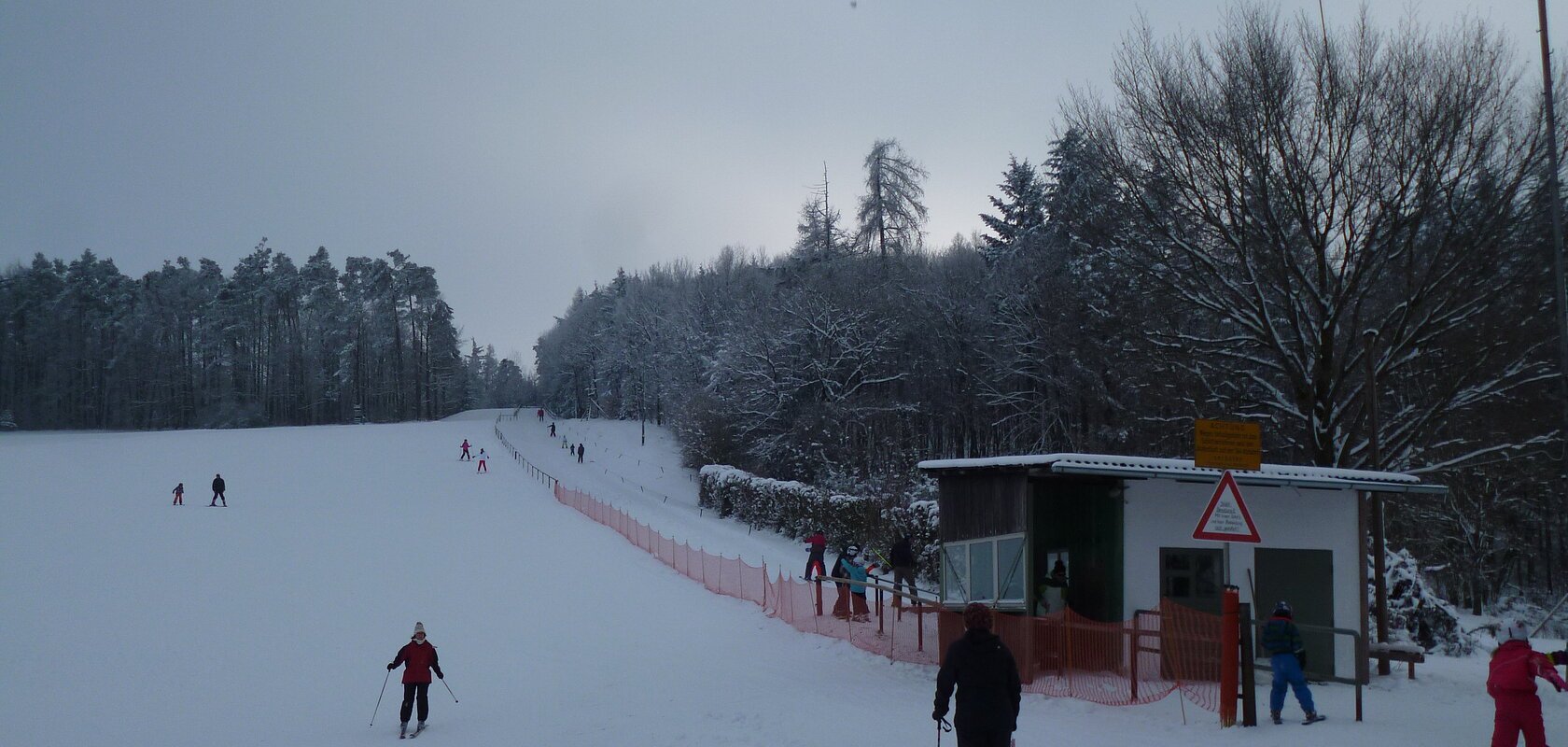 Skifahrer auf einer schneebedeckten Piste neben einem kleinen Schlepplift und einem Wald im Hintergrund.