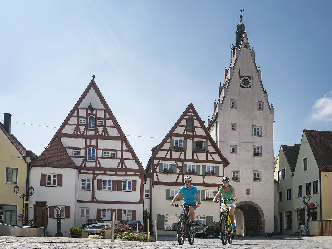 Zwei Personen fahren mit Fahrrädern auf einem gepflasterten Platz, der Monheimer Altstadt. Im Hintergrund Fachwerkhäuser und ein Turm mit einem Torbogen und einer Uhr.