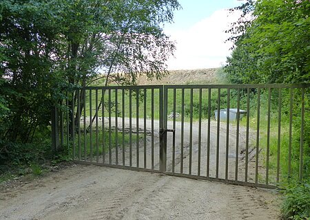 Verschlossenes Metalltor auf einem unbefestigten Weg, umgeben von Bäumen und Gras. Im Hintergrund ist ein Müllcontainer und ein Hügel sichtbar.