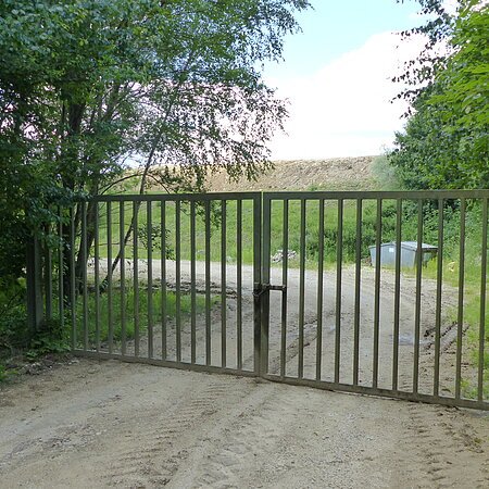 Verschlossenes Metalltor auf einem unbefestigten Weg, umgeben von Bäumen und Gras. Im Hintergrund ist ein Müllcontainer und ein Hügel sichtbar.