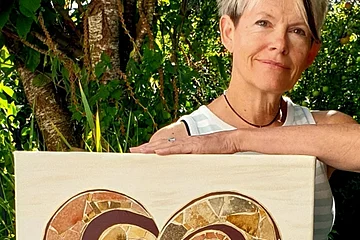 Eine Person hält ein Mosaikbild mit Herzmotiv vor einem Baum im Freien. Steinkunstwerke von Gerlinde Berger