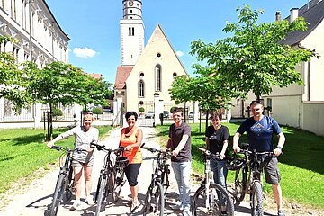 Fünf Personen stehen mit Fahrrädern auf einem Weg vor einer Kirche mit Turm, umgeben von Bäumen und Gebäuden. Im Hintergrund blauer Himmel.