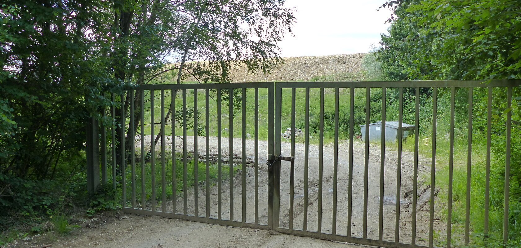 Verschlossenes Metalltor auf einem unbefestigten Weg, umgeben von Bäumen und Gras. Im Hintergrund ist ein Müllcontainer und ein Hügel sichtbar.