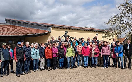 Große Gruppe von Menschen steht vor einem gelben Gebäude mit rotem Dach und einer Statue hinter Ihnen. Im Hintergrund ein Baum auf einer Wiese und bewölkter Himmel.